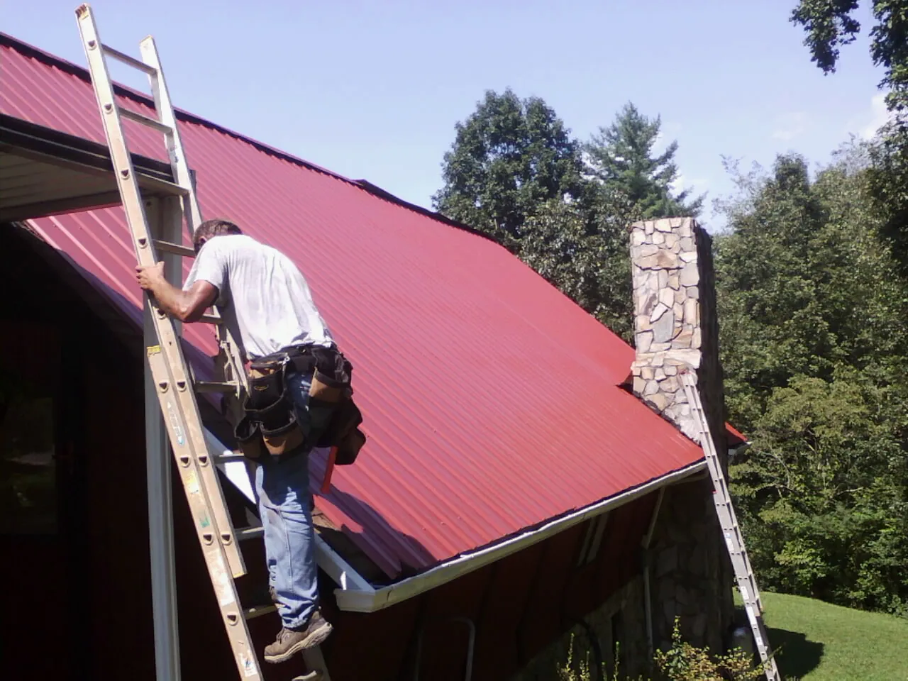 Roofer on a ladder beside a red metal roof and stone chimney — Soffit & Fascia Repair by Roofing Brothers in Redfield